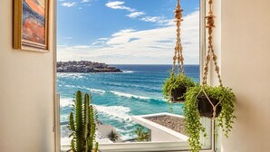 A window with a view of the ocean and a coastal town, two hanging macrame planters with plants, and a framed painting on the wall.