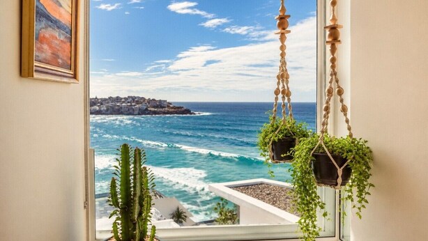 A window with a view of the ocean and a coastal town, two hanging macrame planters with plants, and a framed painting on the wall. - ★ Sub-Penthouse Beach View ★ by Sydney Dreams ★ (Bondi Beach)