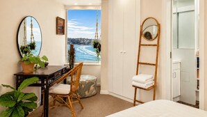 A bedroom with a bed, a desk with a chair, a window with a view of the sea, a hanging plant, and a mirror.