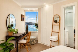 A bedroom with a bed, a desk with a chair, a window with a view of the sea, a hanging plant, and a mirror.