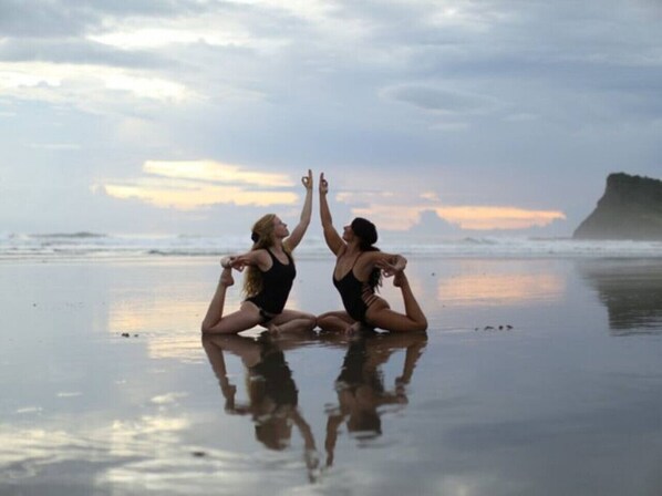 Dos personas realizando una postura de yoga en una playa con un promontorio rocoso al fondo.