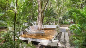 Una terraza de madera con una silla lounge blanca y una pequeña mesa, rodeada de exuberante follaje tropical.