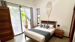 Un dormitorio con cama, una mesita de noche, un atrapasueños en la pared y un ventanal corredero que da a un balcón con plantas.