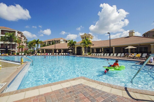 A swimming pool with lounge chairs and palm trees, surrounded by buildings with a Mediterranean architectural style.