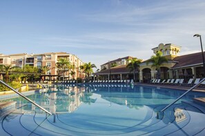 Ein Schwimmbecken mit Liegestühlen und Sonnenschirmen, umgeben von mehrstöckigen Gebäuden mit Balkonen und Palmen.