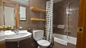 Een badkamer met een wastafel, toilet, bad en douche. Boven het toilet bevinden zich houten planken en er hangt een handdoek aan de muur.