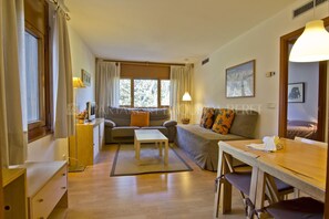 A cozy living room with a sofa, a coffee table, and a dining table. There is a window with curtains, a lamp, and a painting on the wall. - Cabirol - Edificio Bonaigua (Baqueira)