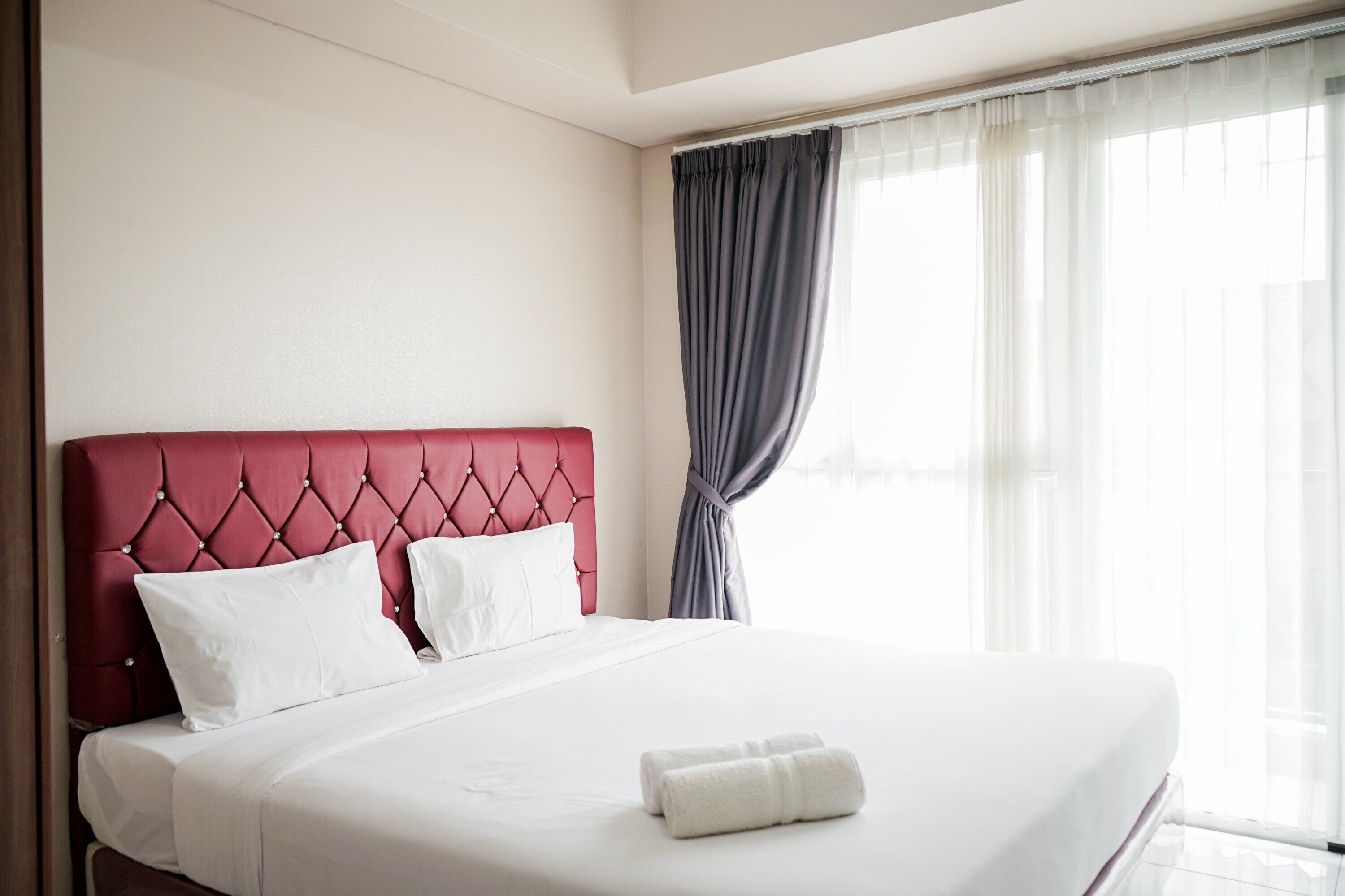 A neatly made bed with white linens and a red headboard, two white pillows, and a rolled white towel on the bed.