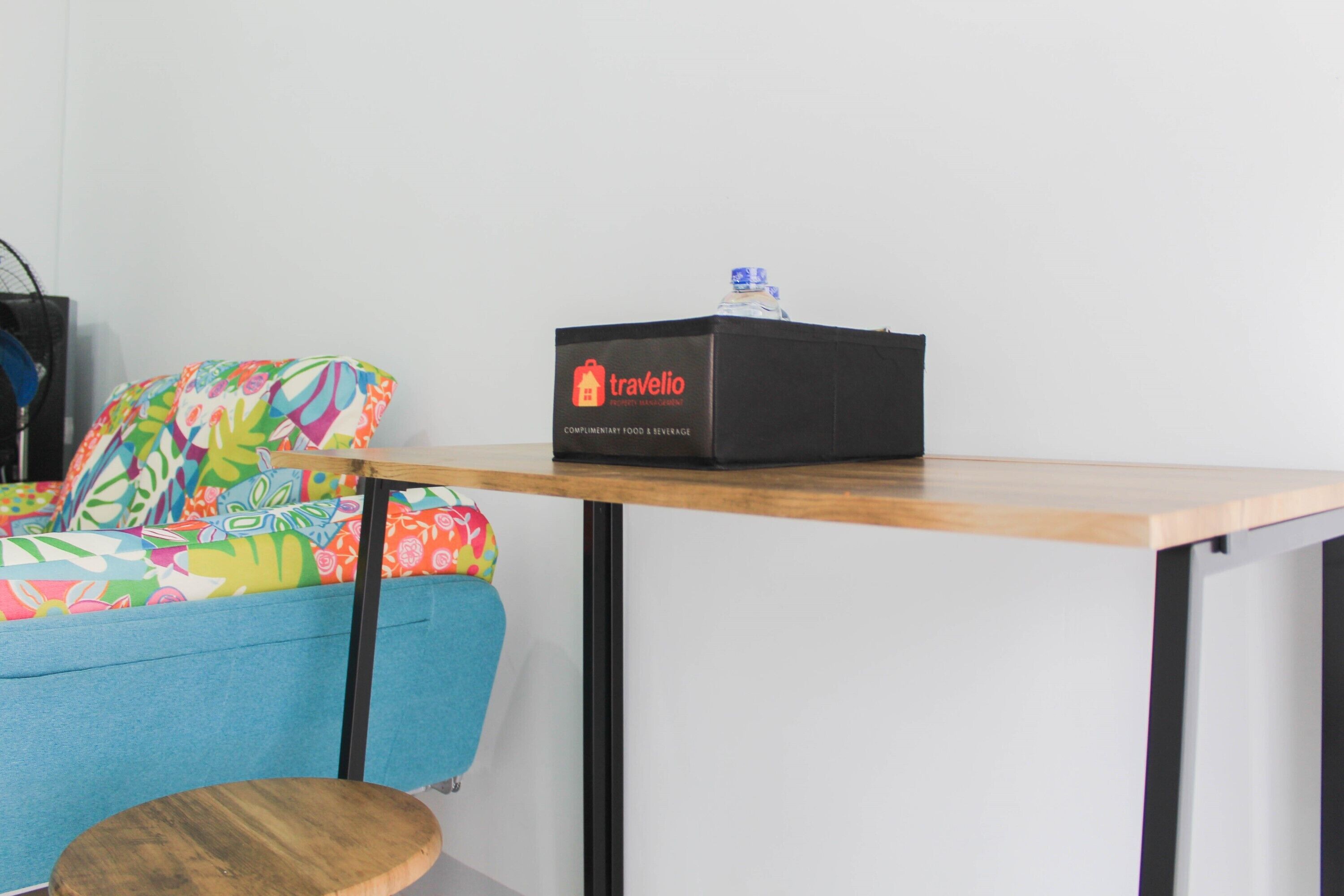 A traveler's room with a colorful sofa, a small wooden stool, and a black box labeled 'Travelio' on a wooden table.