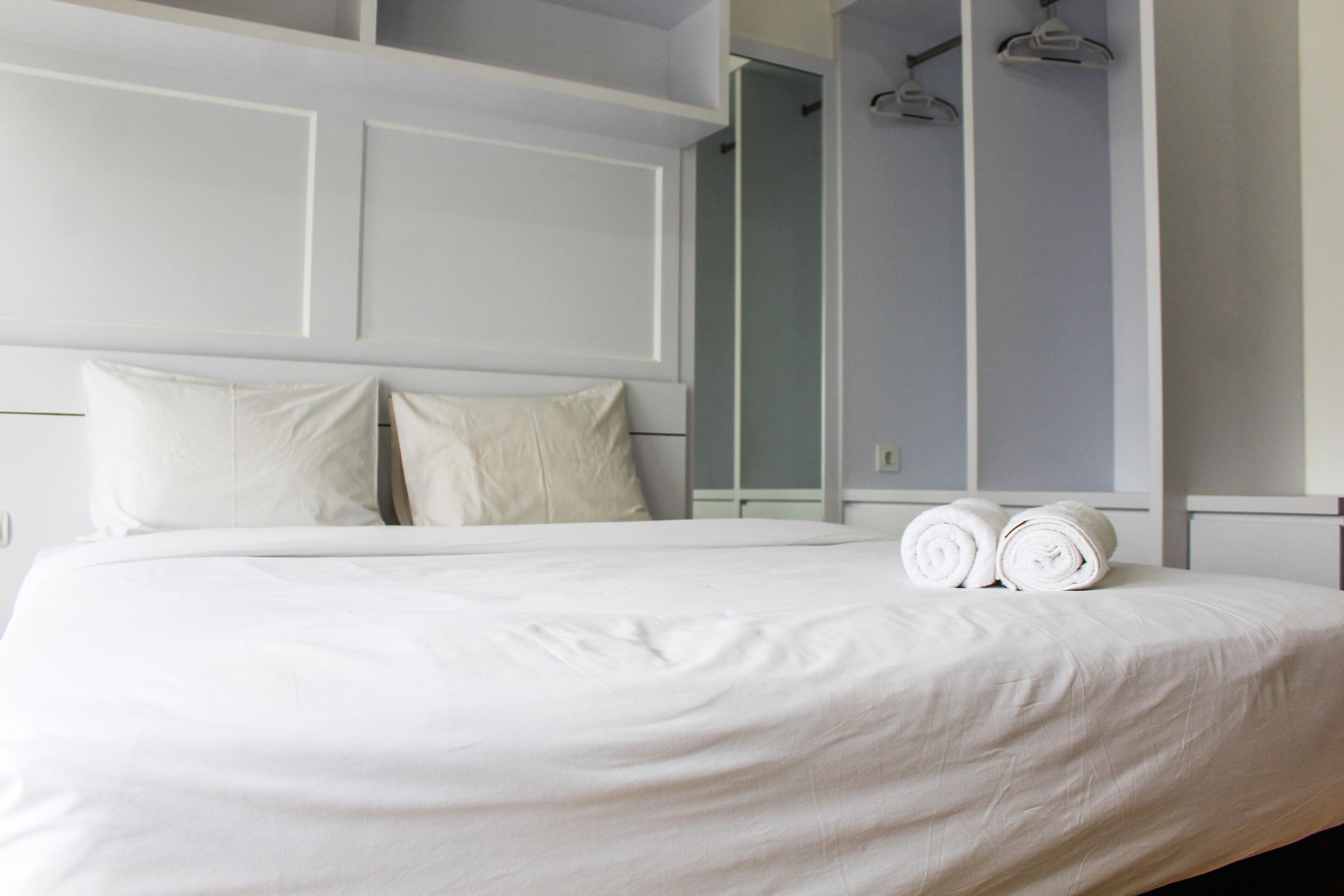A neatly made bed with white linens and two rolled towels, a white headboard, and a mirror on the wall.