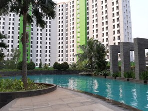 A swimming pool with a clear blue water surface, surrounded by a landscaped area with palm trees and shrubs. In the background, there are high-rise residential buildings with balconies. - Minimalist and Comfy Studio Green Lake View Apartment (South Tangerang)