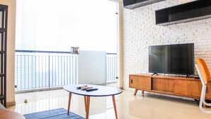 A modern living room with a glass door leading to a balcony, a flat-screen TV, a wooden cabinet, and a small round table. - Homey and Warm 3BR Apartment at Ambassade Residences (Jakarta)