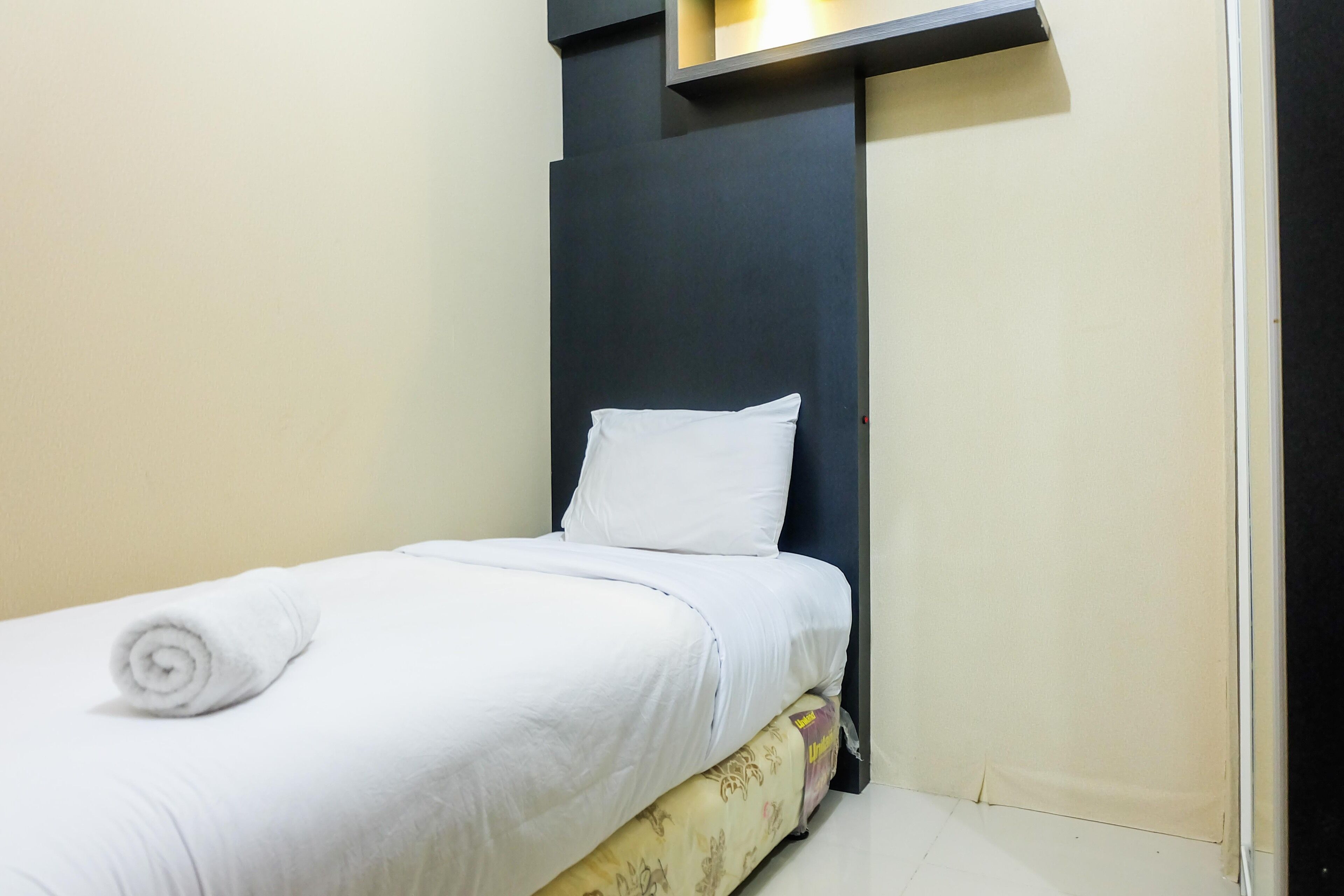 A single bed with white bedding and a rolled towel, a dark headboard, and a small shelf above the bed.