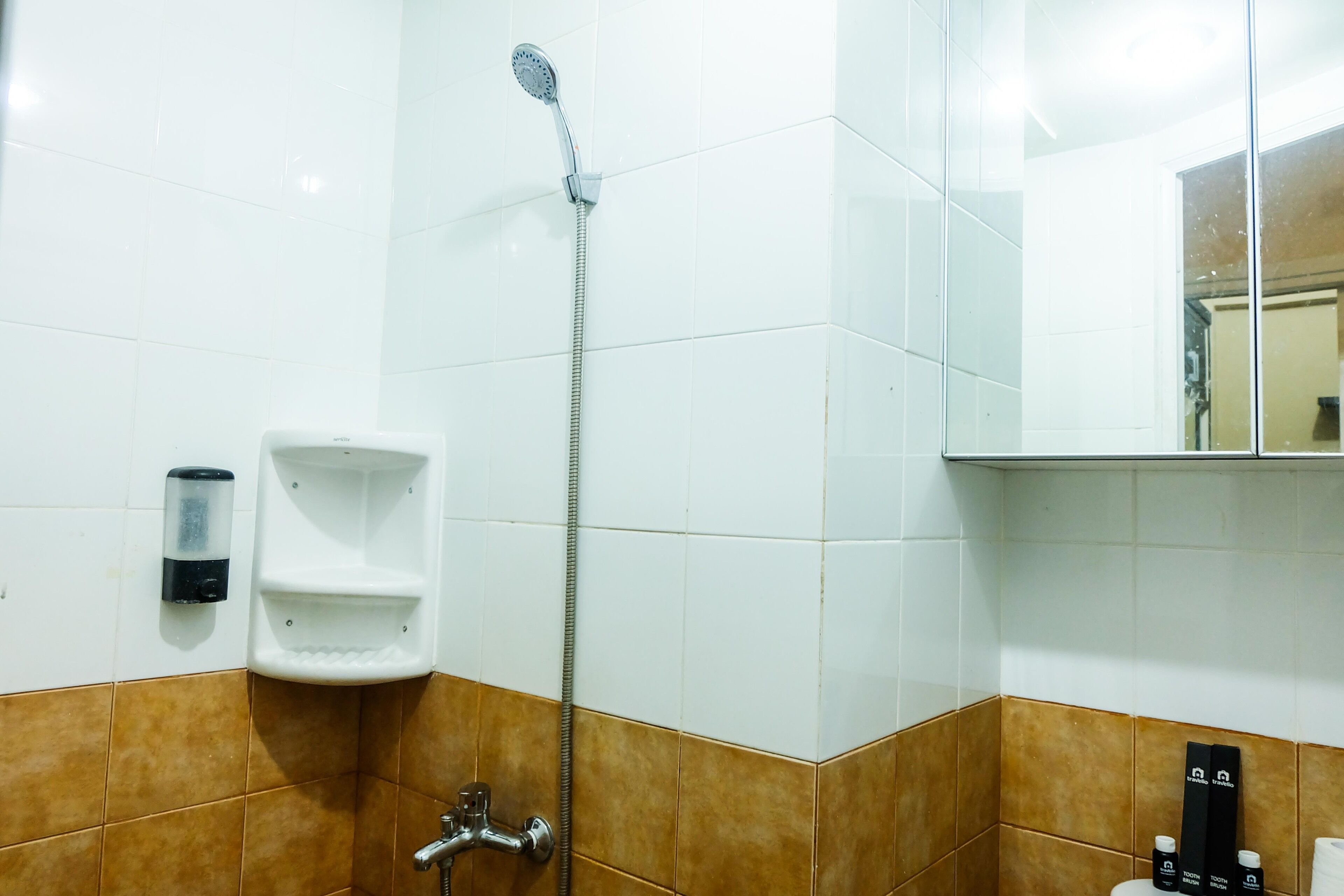 A bathroom with white and brown tiles, a handheld showerhead, a soap dispenser, and a toilet paper holder.