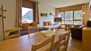 Une pièce chaleureuse avec une table à manger en bois, un canapé et un lit. De grandes fenêtres sont agrémentées de rideaux, et on y trouve également une télévision, ainsi qu’une étagère remplie de livres et d’une guitare.