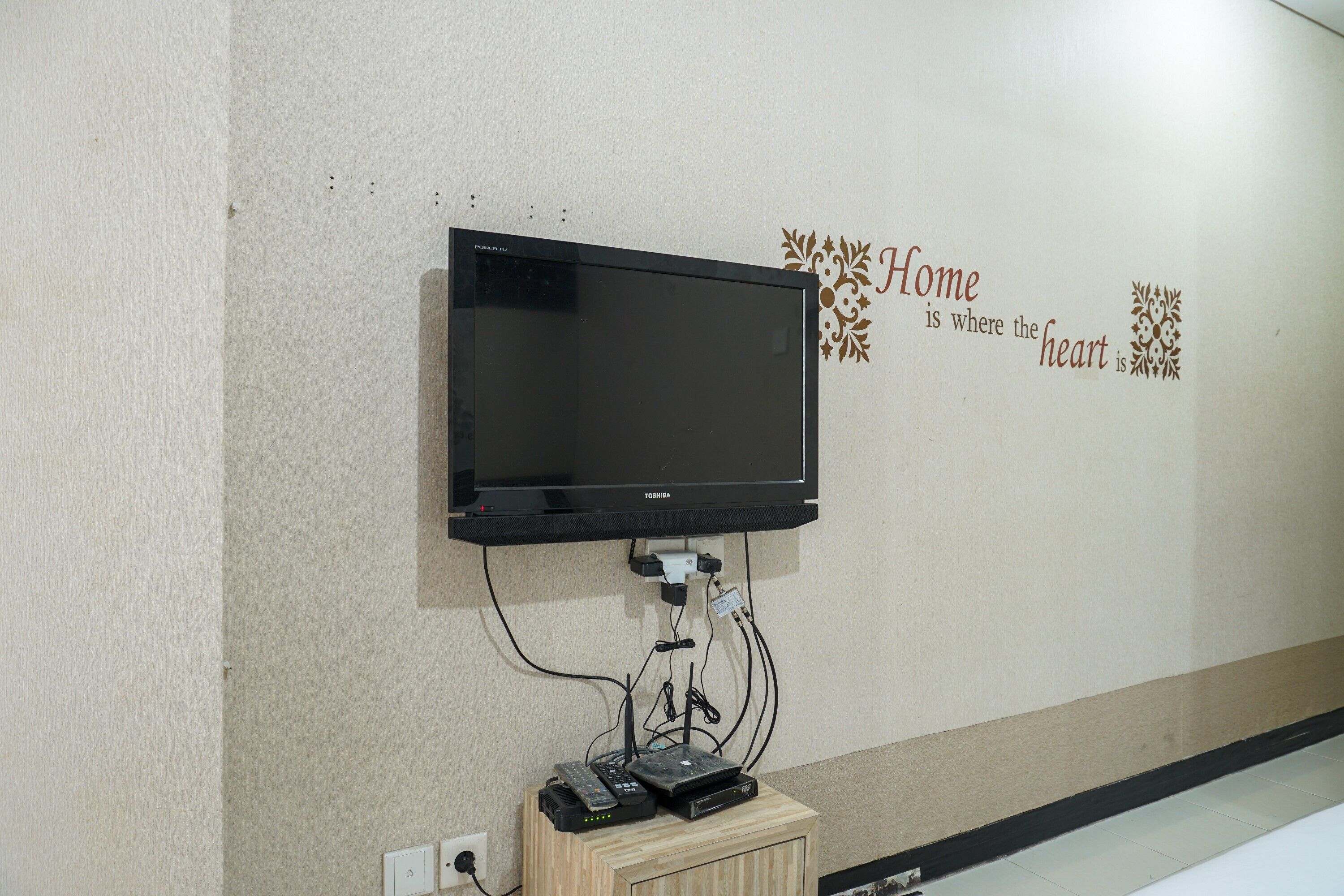A wall-mounted television with a Toshiba logo, a decorative wall sign saying "Home is where the heart is," and a wooden cabinet with electronic devices.