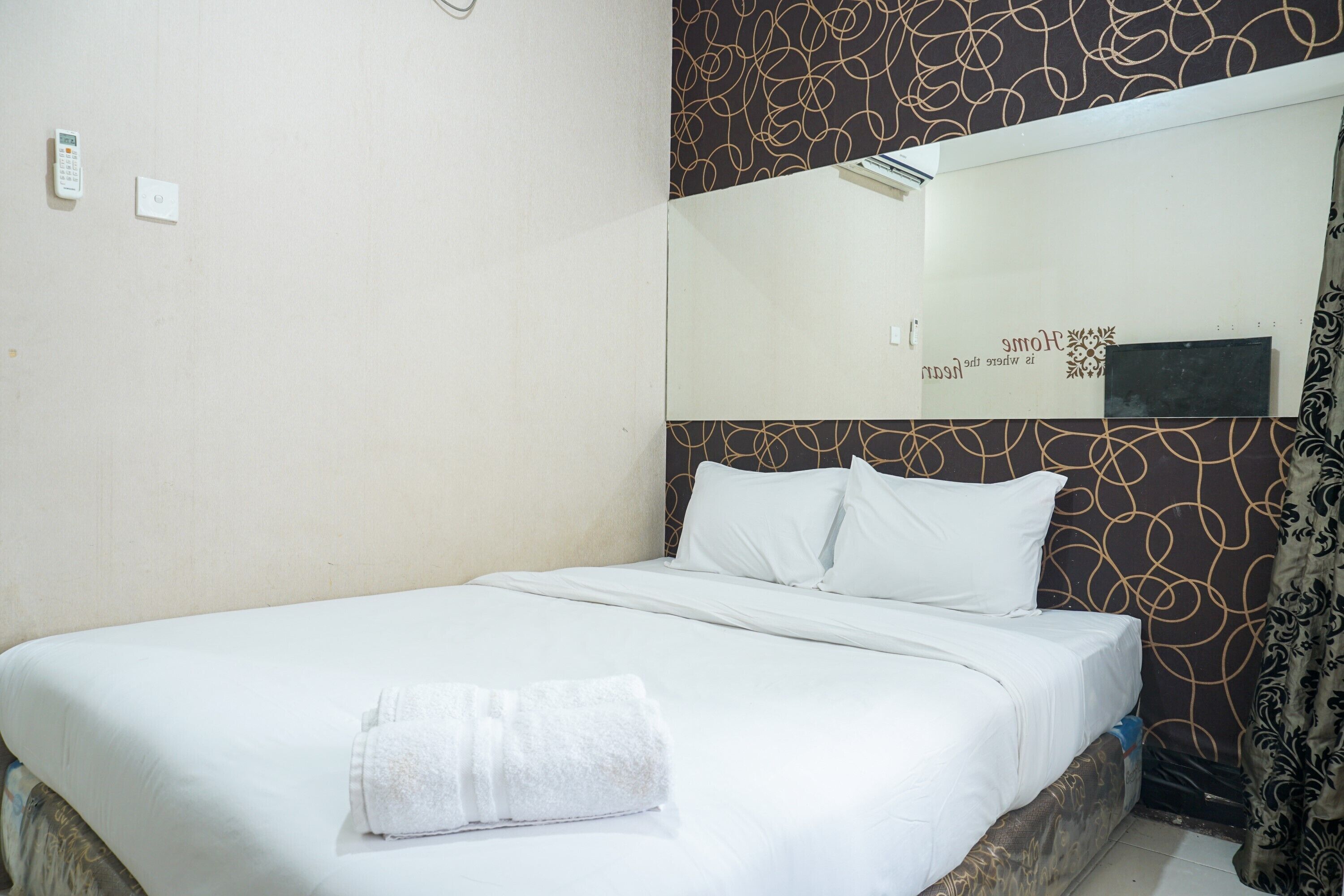 A neatly made bed with white linens and a folded towel, a bedside table, a wall-mounted control panel, and a mirror reflecting part of the room.