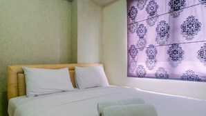 Een net opgemaakt bed met witte lakens en kussens, een opgerolde handdoek op het bed en een raam met een paars bloemenmotief.
