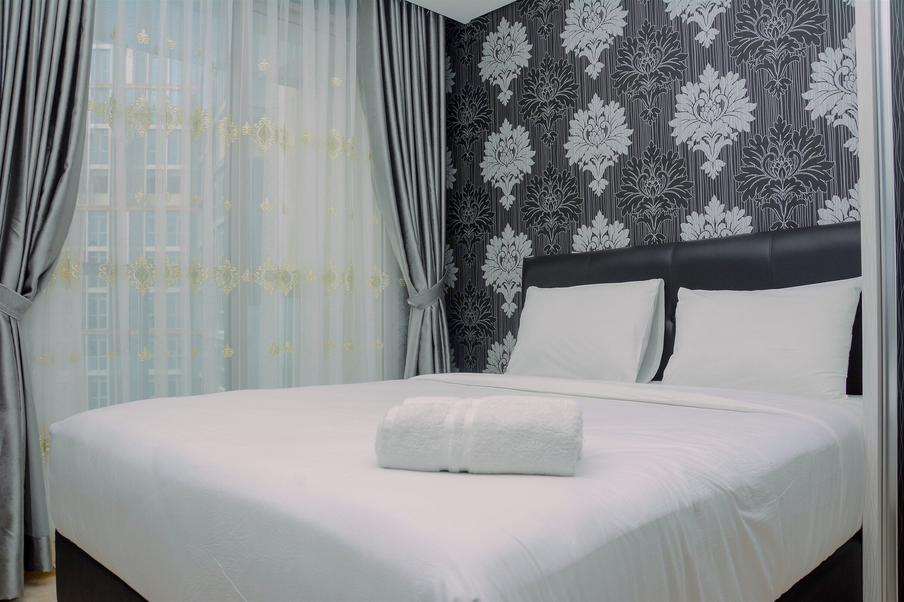 A neatly made bed with a white duvet and a folded white towel, a black headboard, and a patterned wallpaper with floral designs.