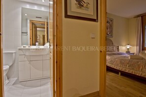Een hotelkamer met een houten vloer, een bed met een beige dekbedovertrek, een nachtkastje met een lamp en een badkamer met toilet en wastafel.