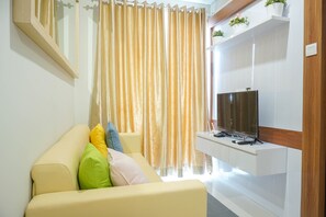 A living room with a beige sofa, colorful cushions, a flat-screen TV on a white stand, and a large window with sheer curtains.