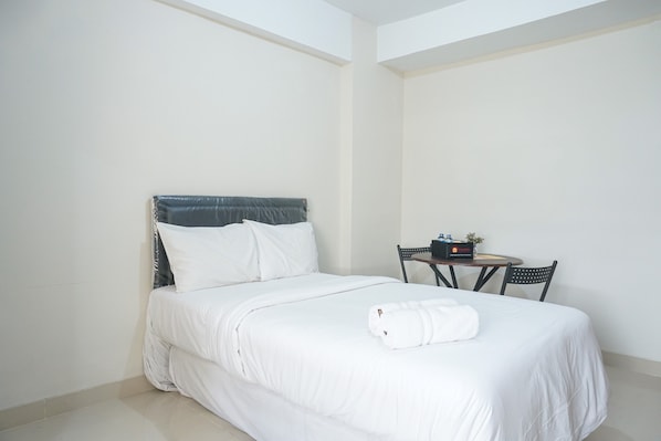 A neatly made bed with white bedding and pillows, a small table with a bottle and a book, and two chairs.