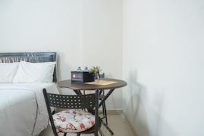 A small, neatly arranged hotel room with a bed, a round table with a chair, a small potted plant, and a box labeled "Travelio."