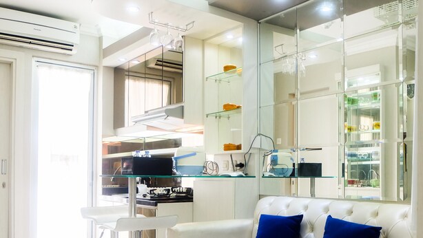 A modern living room with a white sofa, two blue cushions, a glass coffee table, and a kitchen area with a stove and cabinets. - Posh 2BR Apartment at Bassura City near Shopping Mall (Jakarta)
