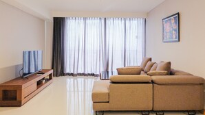A modern living room with a flat-screen TV on a wooden console, a beige sectional sofa, and a large window with sheer curtains. - Big Size with City View @ 3BR Lexington Apartment (Jakarta)