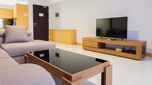 A modern living room with a sofa, a glass coffee table, a TV on a wooden stand, and a door in the background. - Big Size with City View @ 3BR Lexington Apartment (Jakarta)