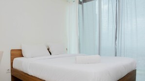 A modern bedroom with a wooden bed frame, white bedding, and a large window with sheer curtains. - Comfy and Warm Studio Grand Kamala Lagoon Apartment (Bekasi)