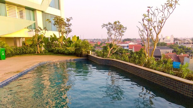A swimming pool with tiled edges, surrounded by greenery and a modern building with large windows. - Comfy and Warm Studio Grand Kamala Lagoon Apartment (Bekasi)