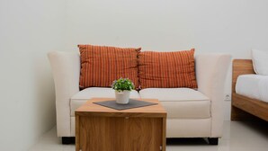 A modern, minimalist room with a beige sofa, two orange-striped pillows, a small wooden side table with a potted plant, and a wooden bed frame. - Comfy and Warm Studio Grand Kamala Lagoon Apartment (Bekasi)