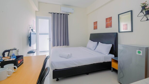 A hotel room with a bed, a desk with a kettle, a refrigerator, and a window with curtains. - Studio Apartment Bogorienze Resort near The Jungle Water Adventure (Bogor)