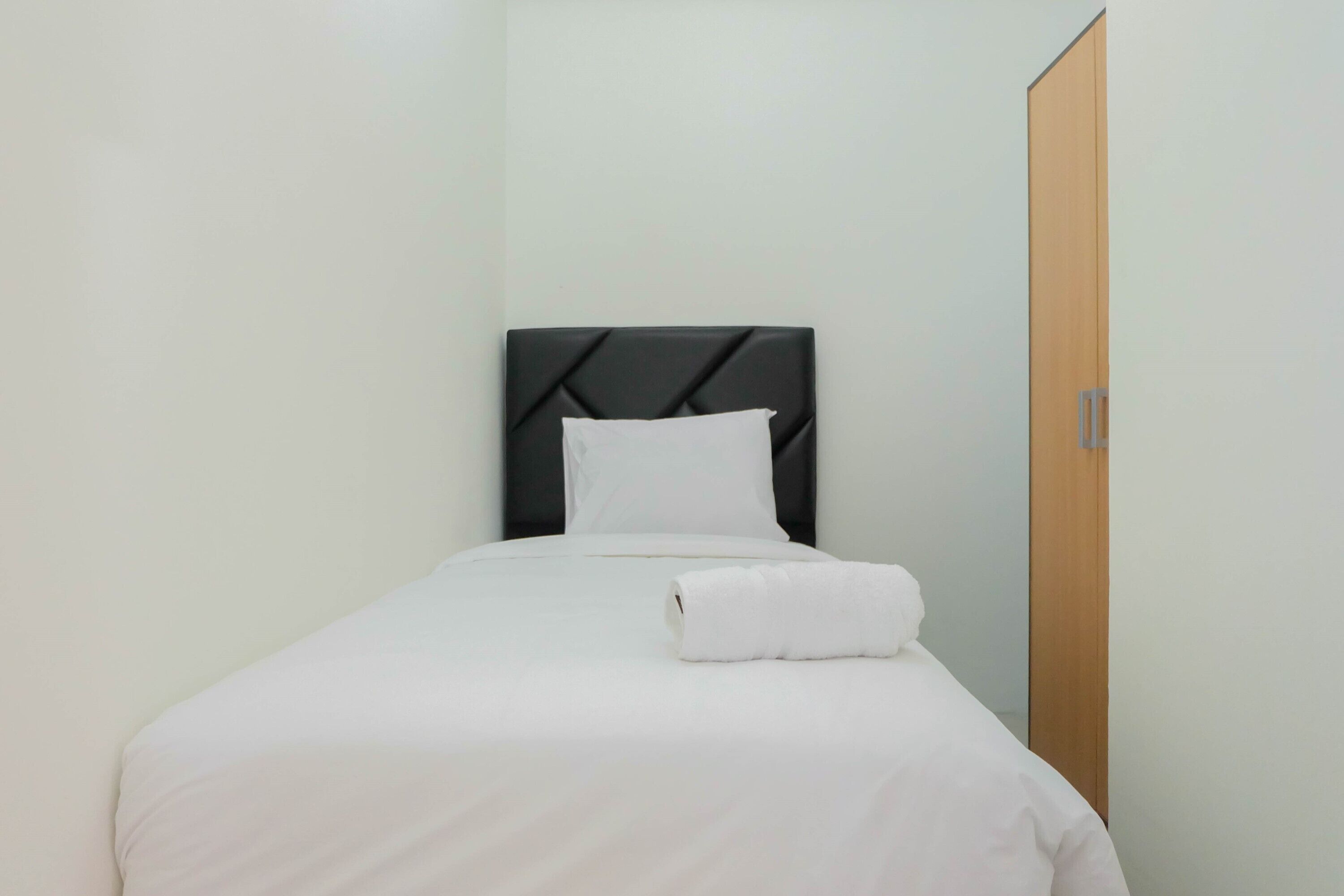A single bed with a black headboard, white bedding, and a rolled white towel on the bed.
