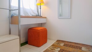 Un dormitorio con cama, mesita de noche con lámpara, un pequeño otoman naranja, una ventana con cortinas traslúcidas y una alfombra estampada en el suelo.