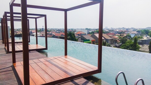 An infinity pool with a wooden deck and metal railings, overlooking a residential area. - Modern and Spacious Studio Grand Kamala Lagoon Apartment (Bekasi)
