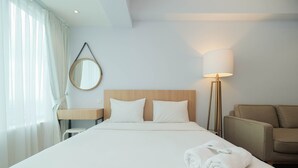 A hotel room with a large bed, a bedside table, a lamp, a sofa, and a mirror on the wall. - Modern and Spacious Studio Grand Kamala Lagoon Apartment (Bekasi)