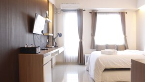 A hotel room with a bed, a desk with a computer, a lamp, and a window with curtains. - Compact Studio Room @ Galeri Ciumbuleuit 2 Apartment (Bandung)