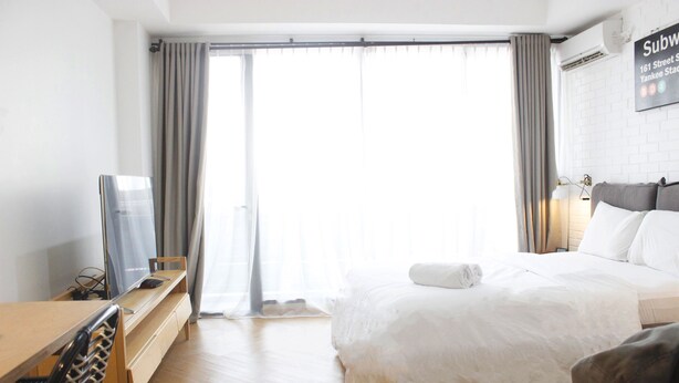 A hotel room with a bed, a desk with a computer, a chair, and a large window with curtains. - Comfy & Artsy Studio at Beverly Dago Apartment (Bandung)