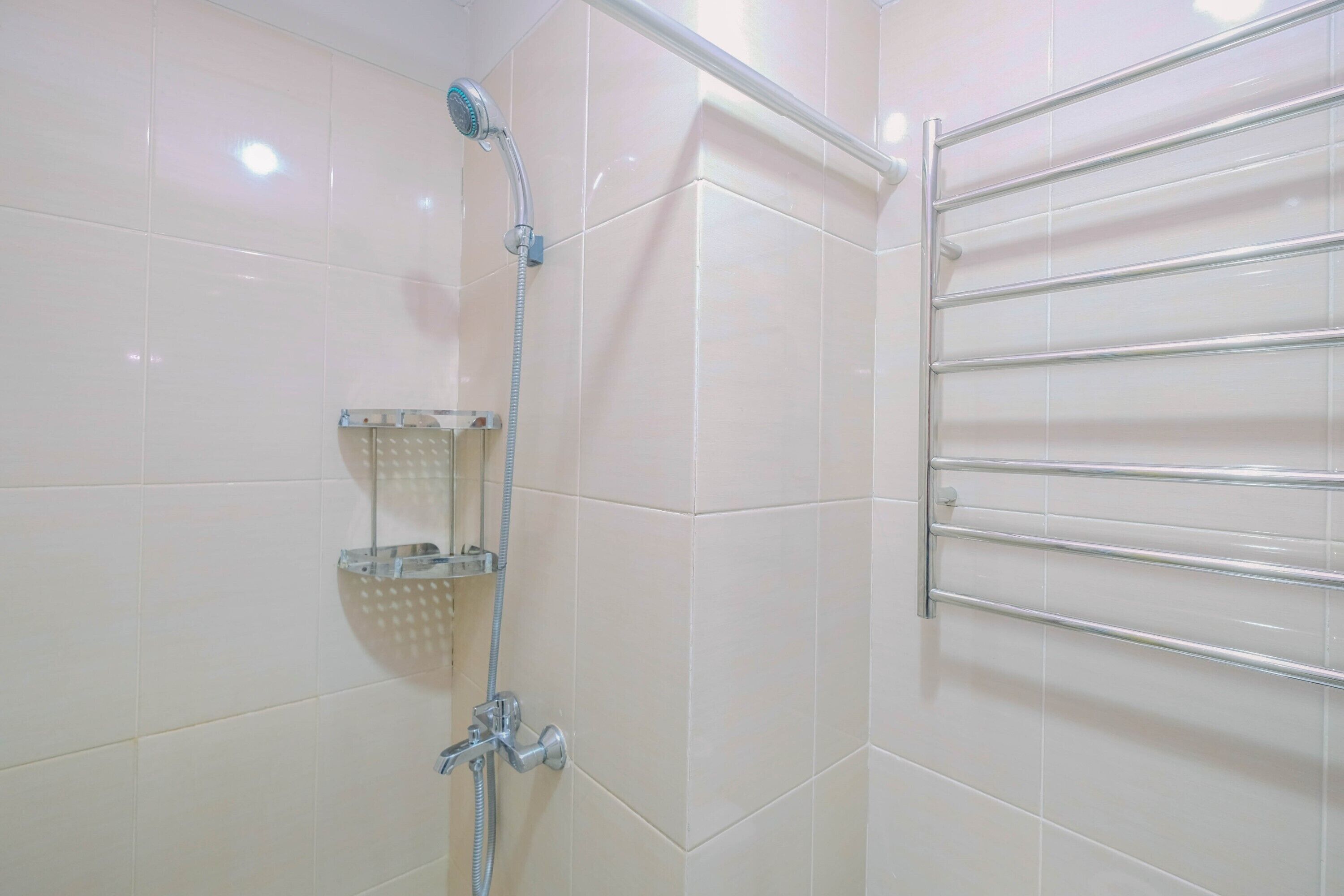 Un baño con azulejos blancos, una ducha con cabezal de ducha manual, un estante de vidrio y un perchero metálico.