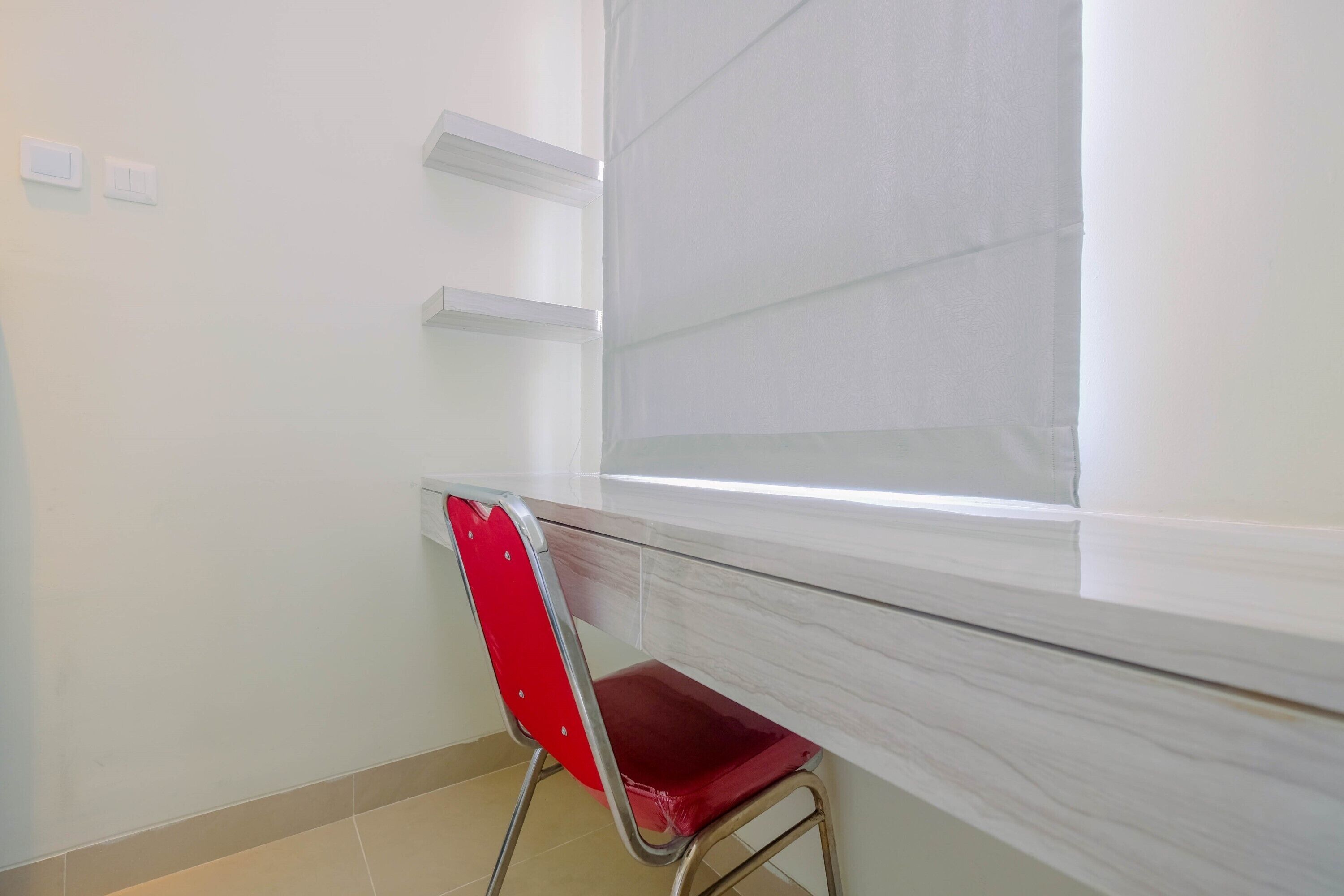 Une pièce avec une chaise rouge, une fenêtre avec des stores et un mur blanc avec un interrupteur.