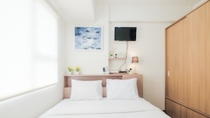 A modern hotel room with a bed, a TV mounted on the wall, a wooden wardrobe, and a small desk with a lamp and decorative items. - Spacious and Comfortable 2BR Cinere Resort Apartment (Depok)