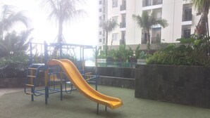 A playground with a yellow slide and climbing structures, surrounded by greenery and a building in the background. - Spacious and Comfortable 2BR Cinere Resort Apartment (Depok)
