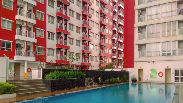 A swimming pool with a tiled edge, surrounded by a building with red and white exterior walls, balconies, and a stone wall with plants. - Comfortable 2BR Apartment at Taman Melati Margonda (Depok)
