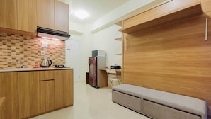 A compact kitchen with wooden cabinets, a tiled backsplash, a kettle, a refrigerator, and a foldable bench. - Modern Studio Apartment 26th on Top of Green Pramuka Mall (Jakarta)