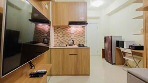 A compact kitchen with wooden cabinets, a tiled backsplash, a built-in stove, a refrigerator, a microwave, and a flat-screen TV. - Modern Studio Apartment 27th on Top of Green Pramuka Mall (Jakarta)