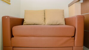 Een bruine leren bank met twee kussens in een kamer met een houten wand en een raam met jaloezieën.