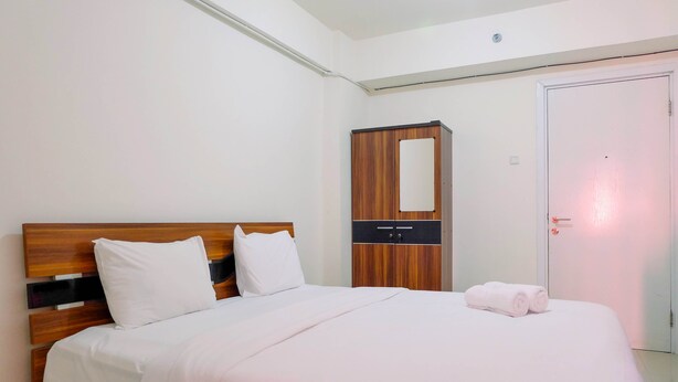 A neatly made bed with white linens and a pink blanket, a wooden wardrobe with a mirror, and a closed door in a simple room. - Simply Studio Apartment at Green Pramuka City (Jakarta)