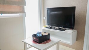 Ein Zimmer mit einem an der weißen Wand befestigten Flachbildfernseher, einem kleinen weißen Tisch mit einer schwarzen Box, die Flaschen enthält, und einer roten Matte, sowie ein Fenster mit Vorhang.